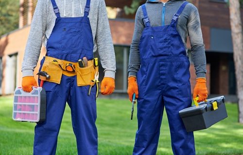 Cropped shot of two male builders in blue overalls carrying toolbox at construction site. Building, protective gear and people concept